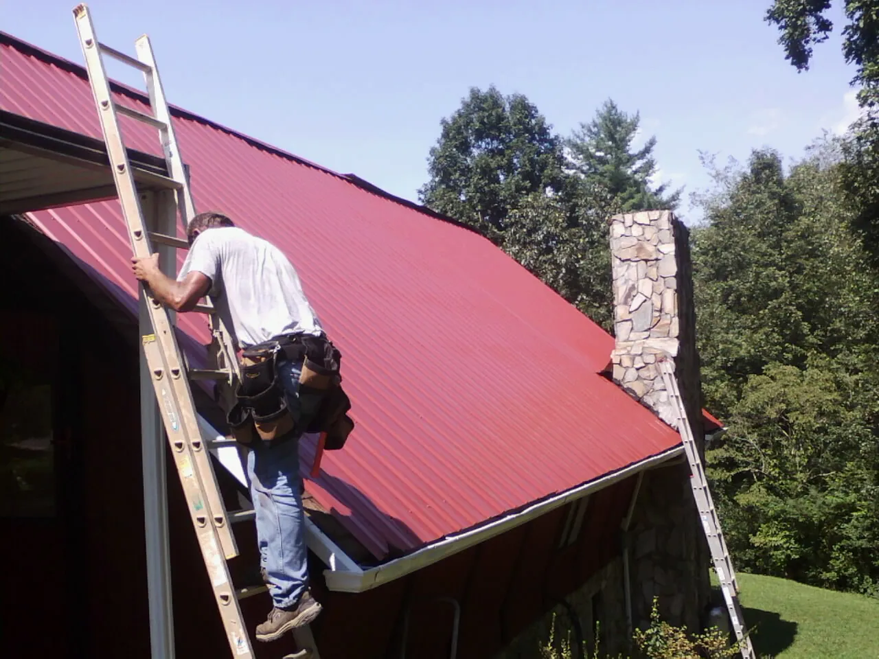 Roofer on a ladder beside a red metal roof and stone chimney — Soffit & Fascia Repair by Roofing Brothers in Catlett